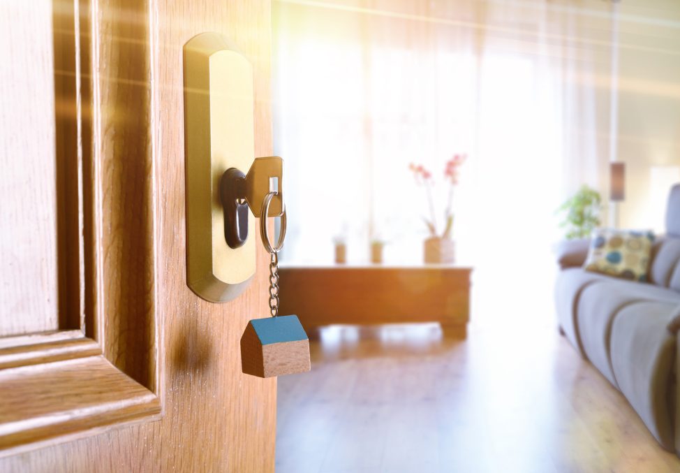Open entrance door detail of a house with keys in the lock and furnished living room in the background with golden light effect.